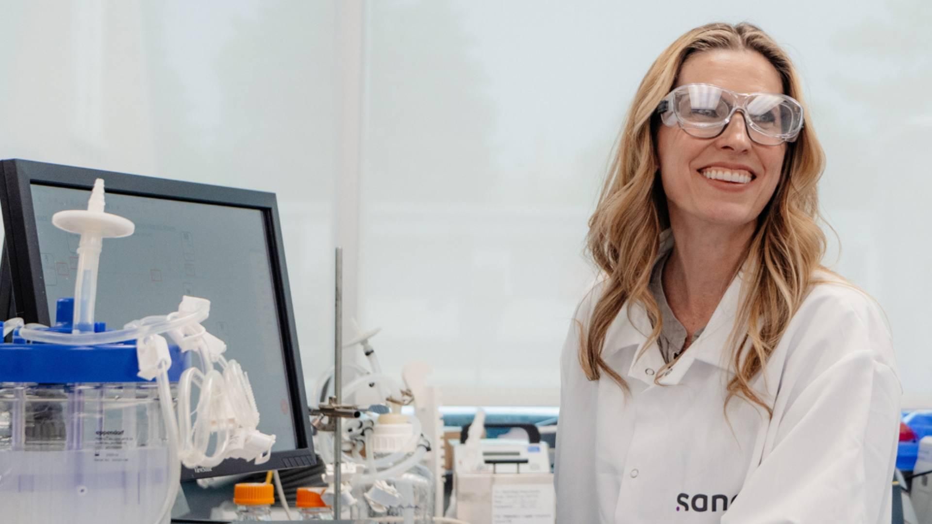 Female, Senior Unit Head, Drug Product Development, wearing a white lab coat and safety glasses and purple gloves, smiles while working at her lab computer